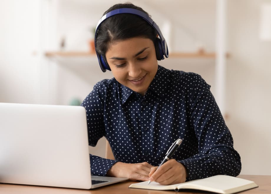 Imagem de uma mulher estudando diante de um notebook com headfones e fazendo anotações